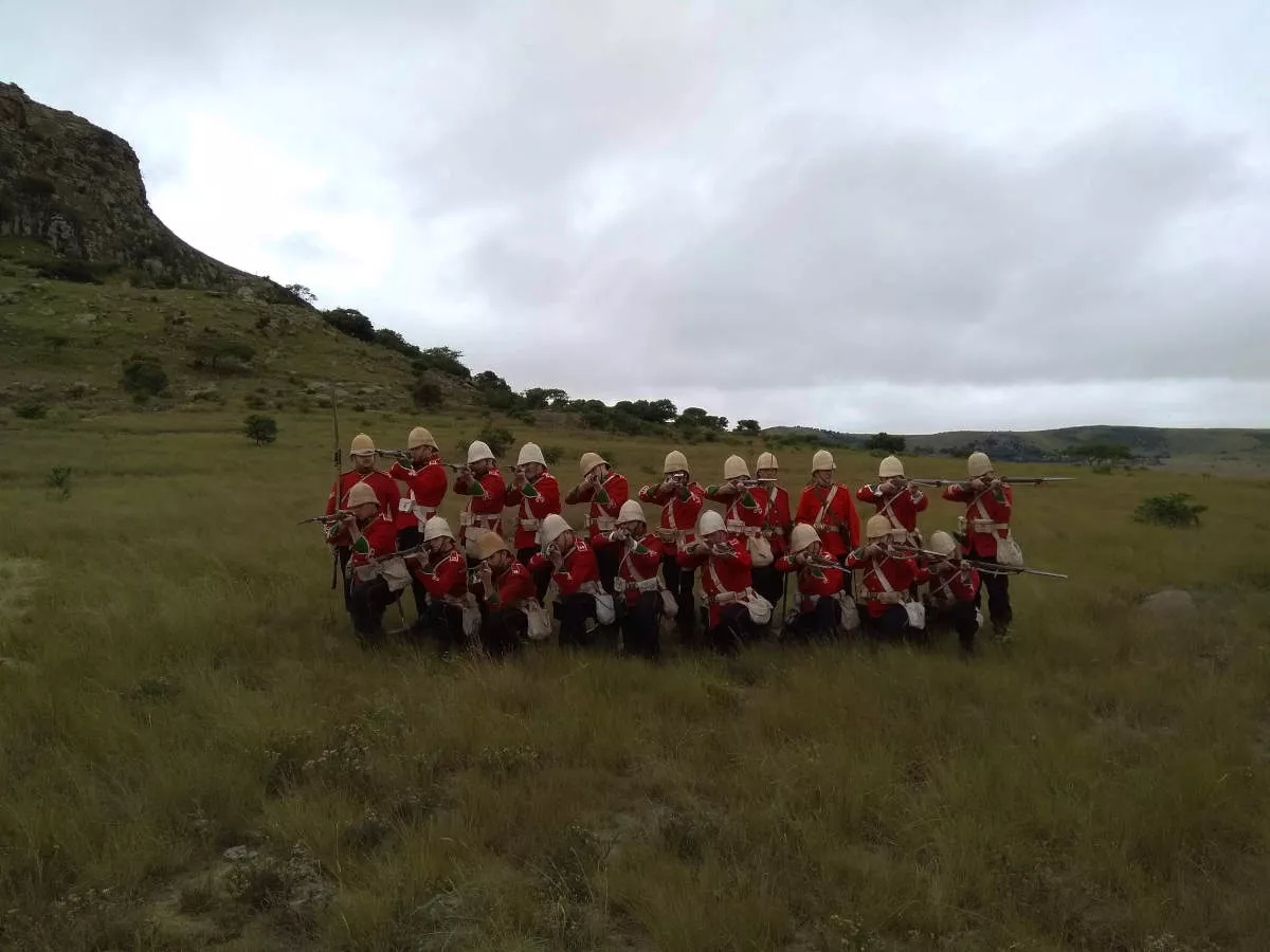 A group fo 20 Britsh reenactors dreesed in 1879's military uniform stand in a company and pose before the hill called isandlwana and point their martini henry rifles at the pphotographer