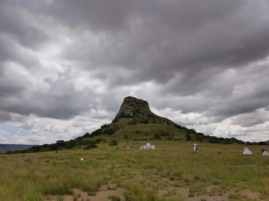 A steep hill called isandlwana is surrounded by dark clouds in the afternoon sky. In the foreground lay piles of stones painted white to mark the remains of fallen british troops.