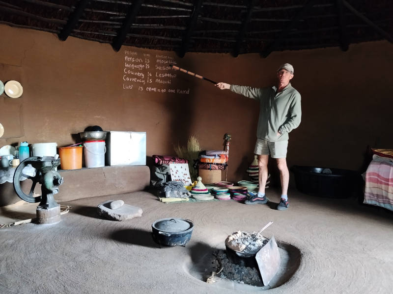A male tour guide stands inside a rural hut in Lesotho and points with a stick to writing on the wall that lists the local currency and has Basotho turns of phrase.